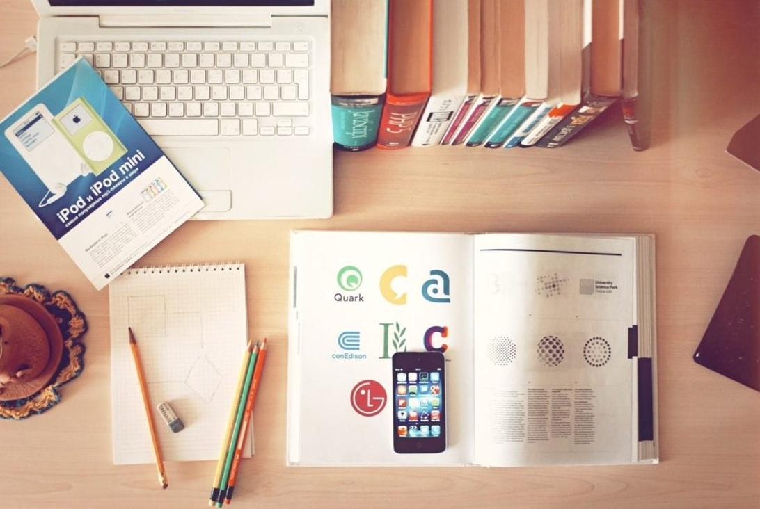 A desk workspace featuring a white laptop, stacked books, a paper notebook with pencils, an iPod manual, and other analog study materials arranged on a wooden surface. This represents a traditional workspace without digital AI elements.